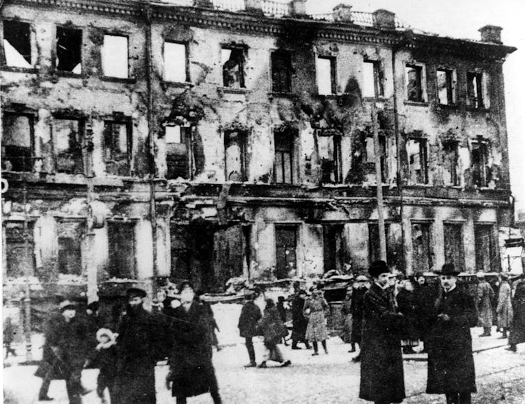 People in long, dark coats during the winter in front of the ruins of a multi-story building.