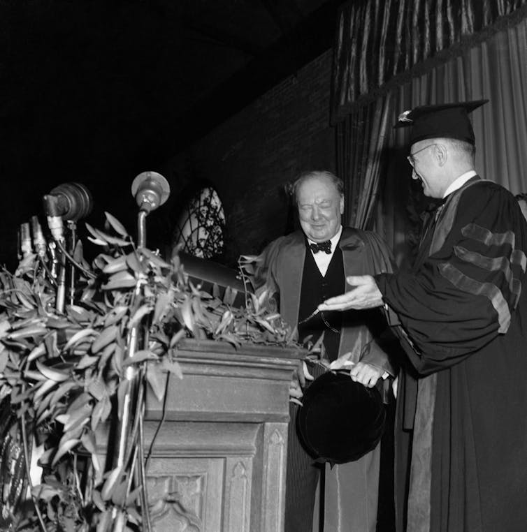 A man in academic robe and cap introduces a chubby man wearing a bow tie as he approaches a lectern.