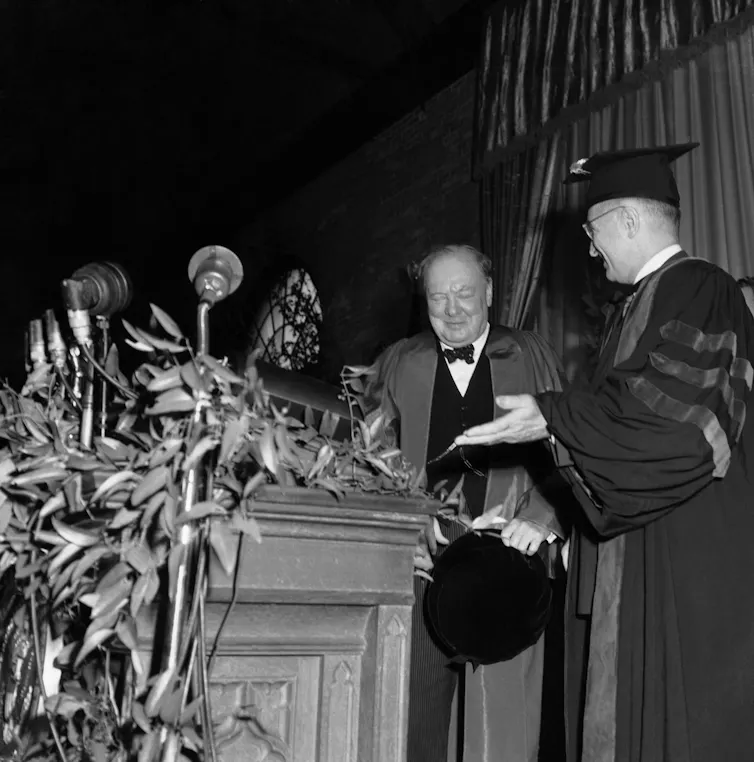 A man in academic robe and cap introduces a chubby man wearing a bow tie as he approaches a lectern.