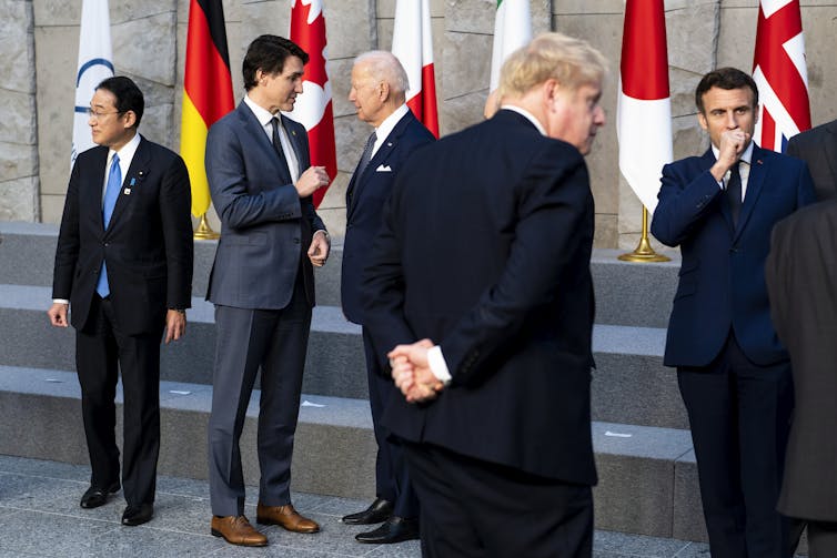 Five men in dark suits standing and talking in front of a flag display.