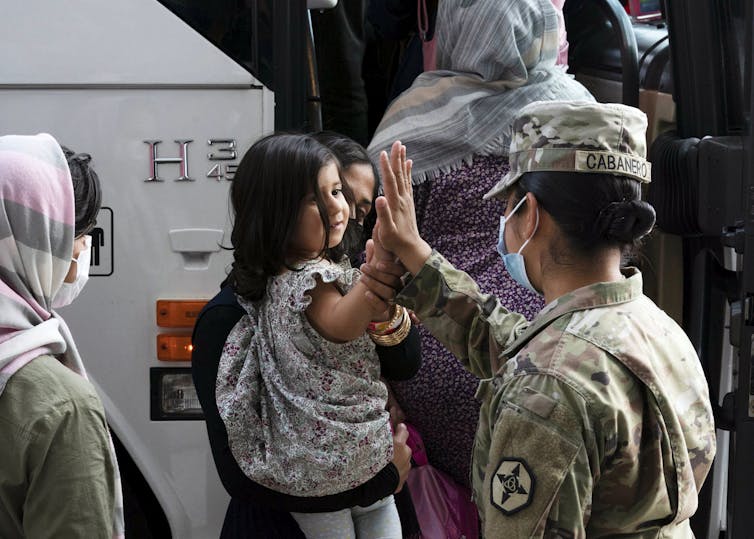 An army officer high-fives a girl carried by a woman outside the door of a bus.
