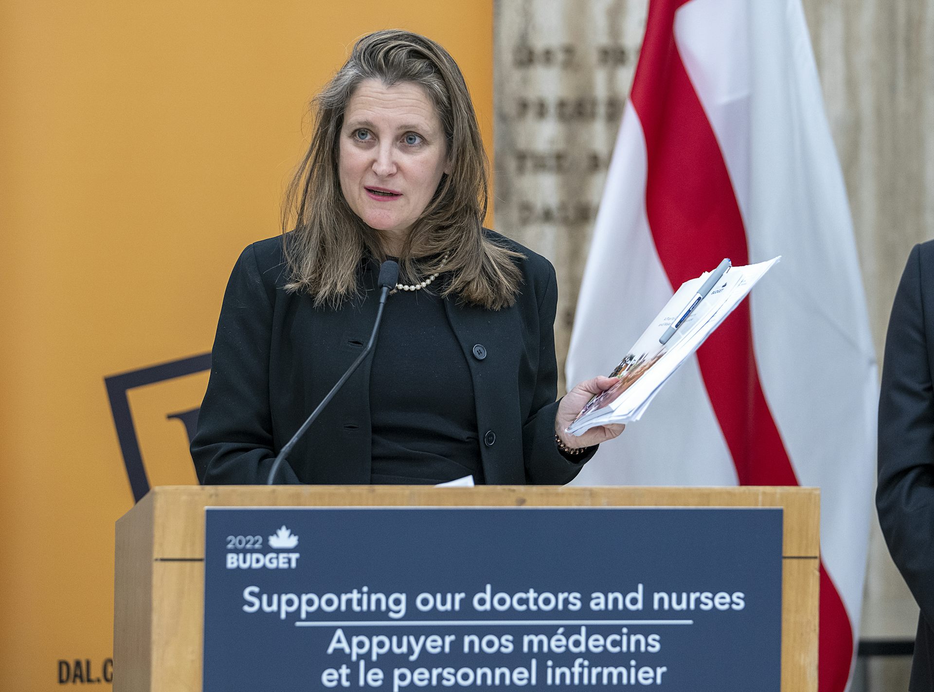 A woman standing and speaking behind a podium. She holds a bundle of papers in one hand.