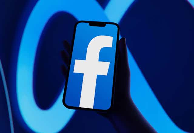 Facebook logo on a phone, held in front of a larger background with Meta's logo resembling an infinity loop