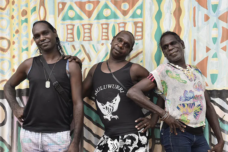 Three Aboriginal transgender sistagirls pose in front of a mural.