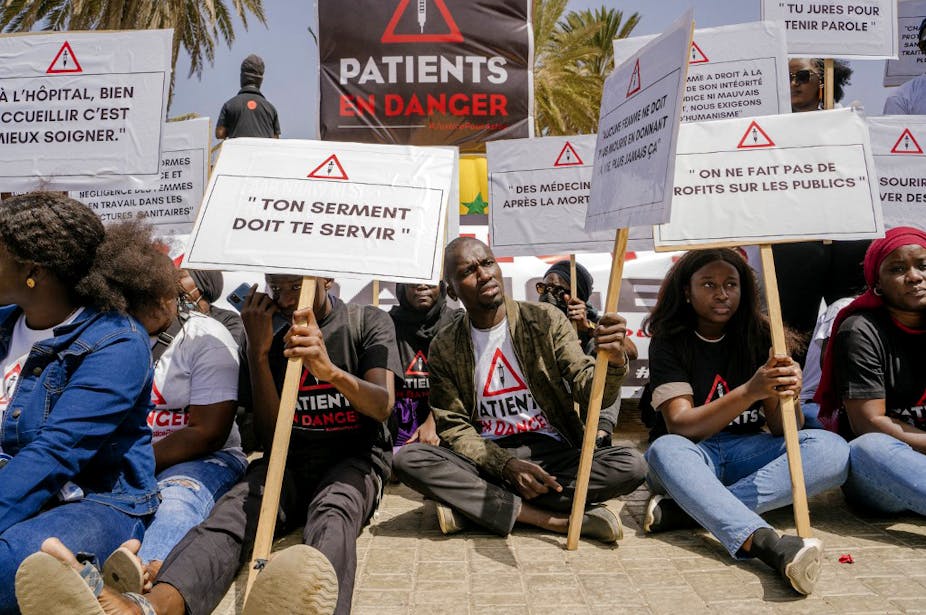 Manifestants assis devant l'hôpital de Dakar, avec des pancartes (Hôpital ; accueillir c'est bien, soigner c'est mieux ; Patients en danger, etc.)