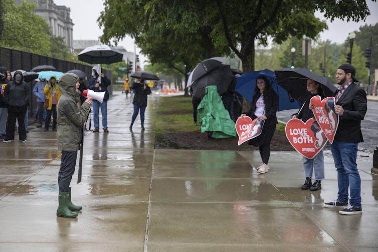 A woman in a hooded jacket using a bullhorn to speak to several other people across the street from her who are holding signs.