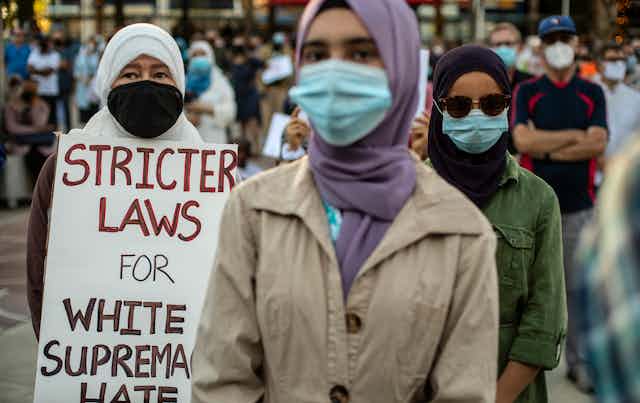 Woman wearing a purple headscarf is in the foreground. Behind her is a women wearing white headscarf and holding a placard that says, "Stricter laws for white supremacy hate".