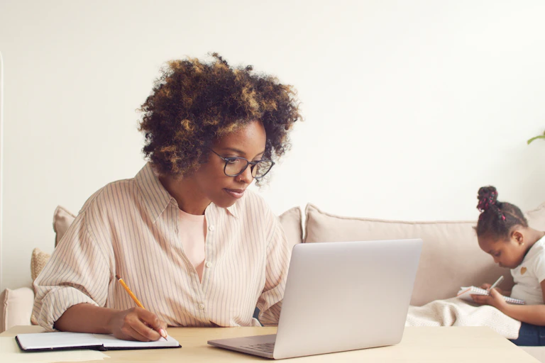 A woman works on a laptop with a child on the couch behind her.