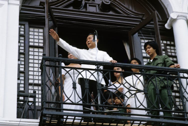 Ferdinand Marcos, dressed in white traditional Philippine shirt, raises his hand and speaks into a microphone to supporters; beside him in a green jumpsuit is his son, Bongbong.