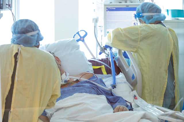 Two nurses in yellow robes and other protective head and face coverings tend to a patient in a hospital bed.