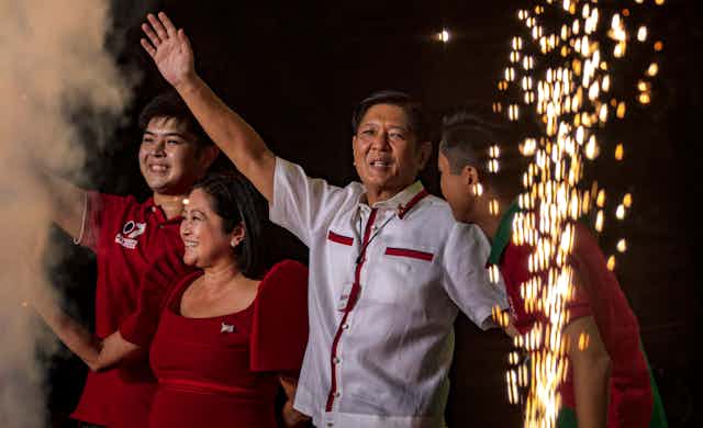 Dressed in a white, traditional Filipino shirt, Bongbong Marcos waves to supporters while surrounded by family members and fireworks.