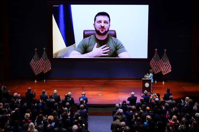 President Zelenskyy wearing a green t-shirt looks out of a large screen on the stage and puts his hand on his heart while the US Congress watches