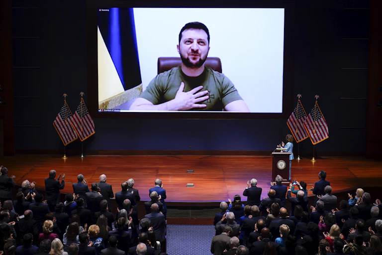 President Zelenskyy wearing a green t-shirt looks out of a large screen on the stage and puts his hand on his heart while the US Congress watches