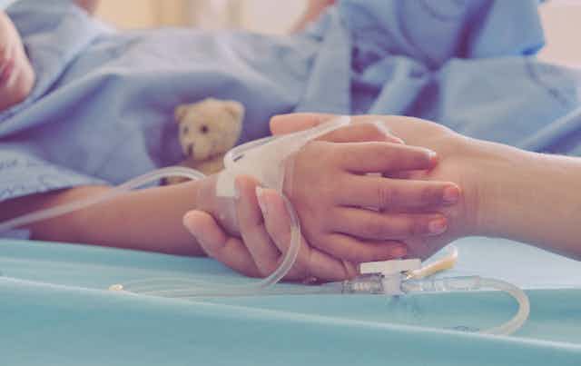 Une adulte tient la main d’un enfant allongé sur un lit d’hôpital, avec un drain.