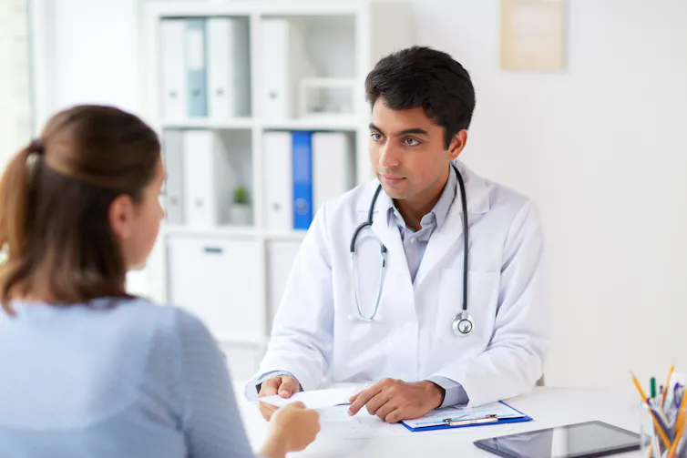 A man in a white coat with a stethoscope around his neck handing a piece of paper to a woman seen from behind