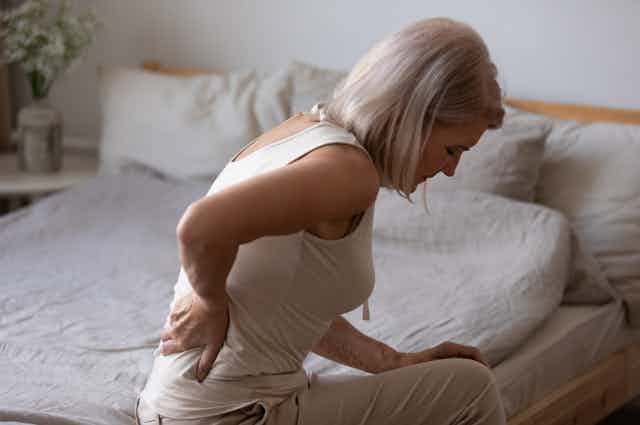 Woman sits on the edge of her bed, bent over in pain, holding her back.
