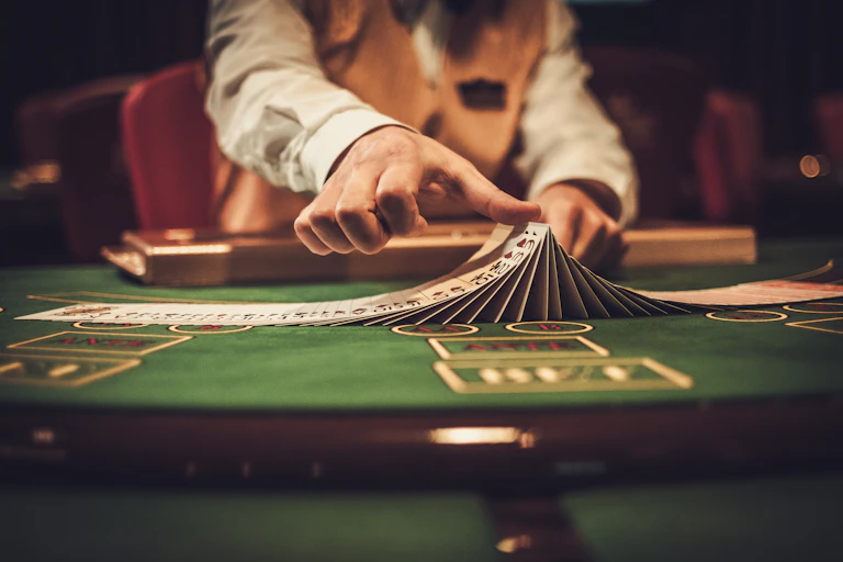 The hands of a croupier wearing a white shirt and orange vest shuffles a deck of cards on a green card table