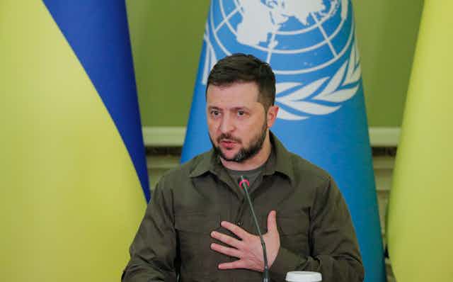 Volodymyr Zelensky speaks into a microphone. He is wearing a green military style shirt and has his hand on his chest. Behind him are Ukrainian and UN flags.