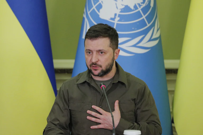 Volodymyr Zelensky speaks into a microphone. He is wearing a green military style shirt and has his hand on his chest. Behind him are Ukrainian and UN flags.