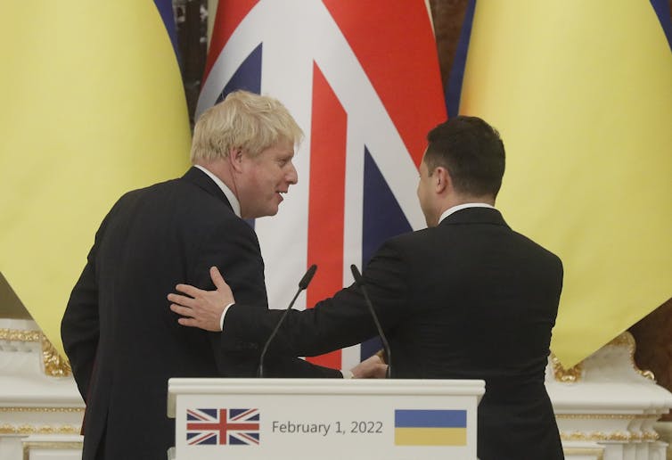 Volodymyr Zelensky claps the UK prime minister, Boris Johnson, on the back while the pair are turned away from a speaking podium. Ukrainian and UK flags are behind them.