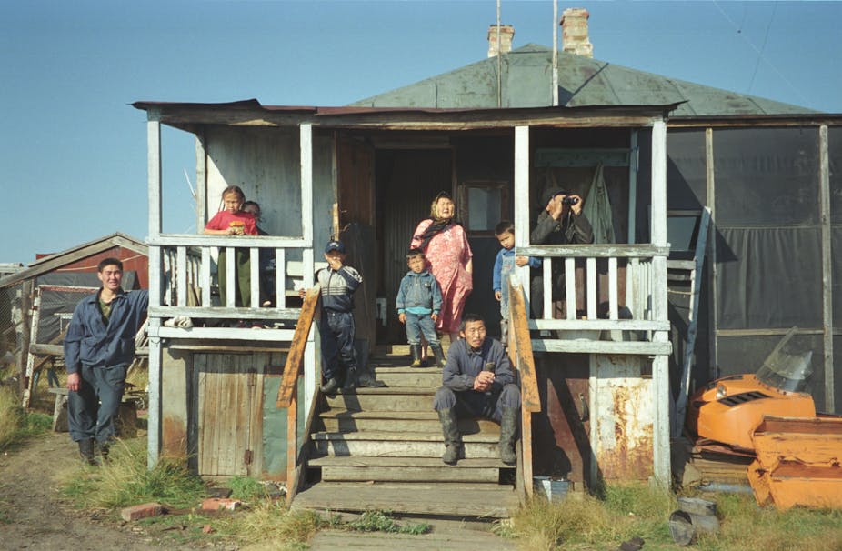 About half a dozen members of a family sit on the porch of a simple wooden house, watching something off camera.