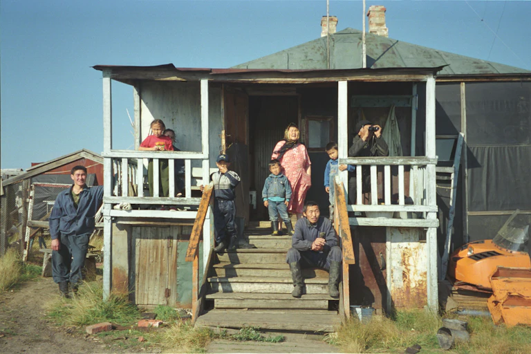 About half a dozen members of a family sit on the porch of a simple wooden house, watching something off camera.
