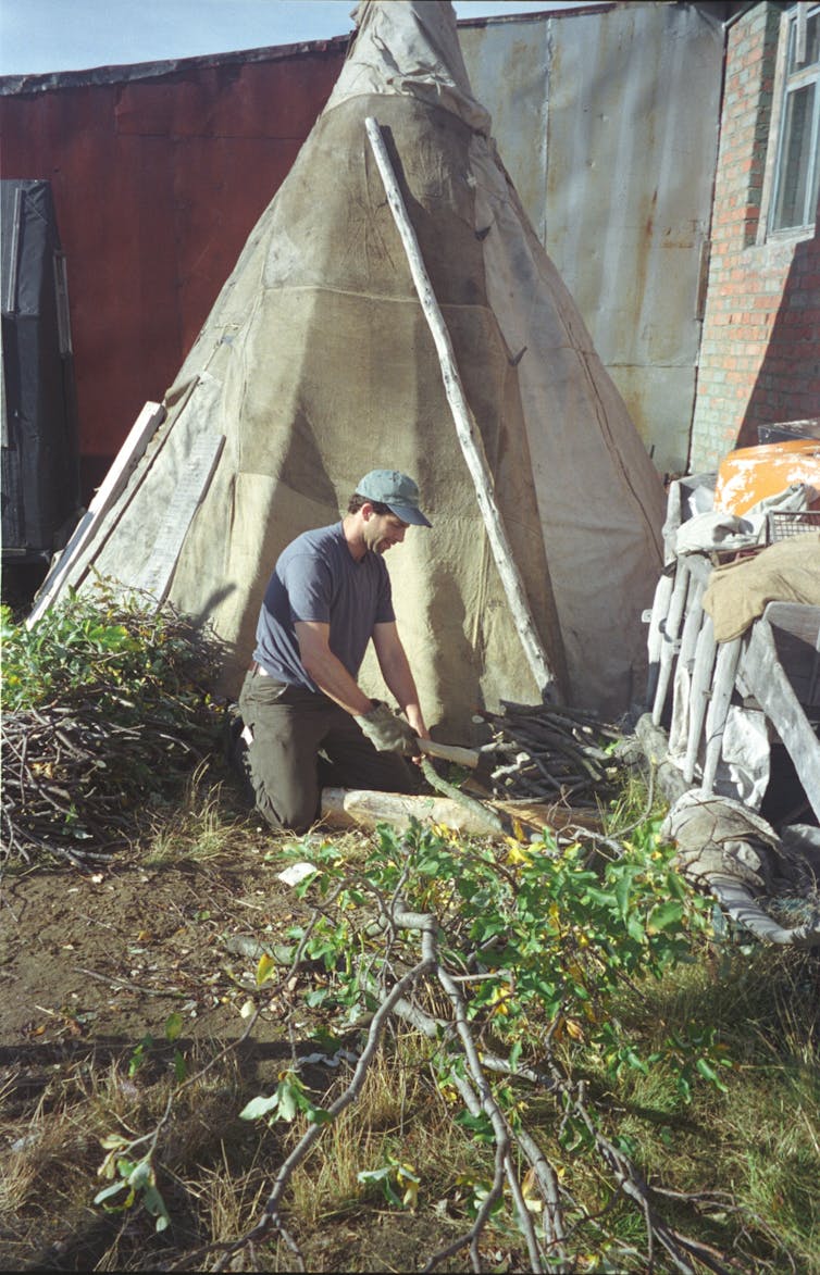 A man in a hat kneels in front of a tent as he chops up small pieces of wood.