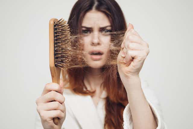 A woman pulls hair out of a hairbrush, appearing dismayed.