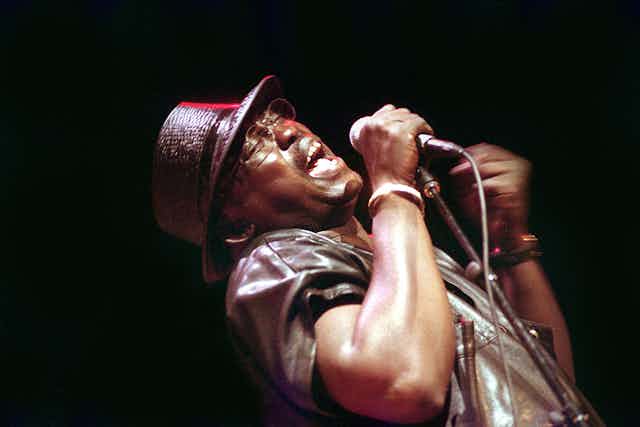 A man in a hat and glasses leans back, eyes shut, singing with a wide open mouth into a microphone he holds in one hand, under a spotlight.