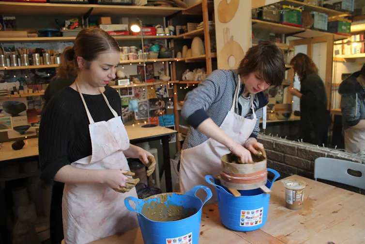 Two people wearing aprons work with clay in buckets on a worktop.