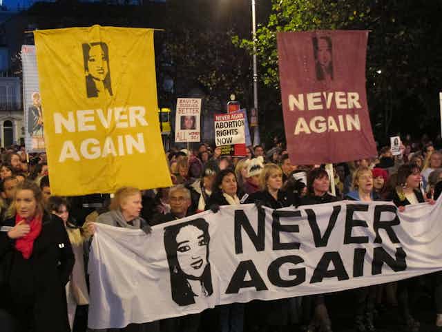 A crowd marches holding signs saying 'never again' and photos of a woman.