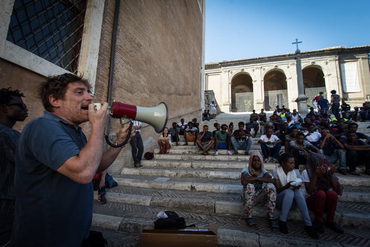 A man in a blue shirt speaks into a voice amplifier while rows of people sit behind him on steps