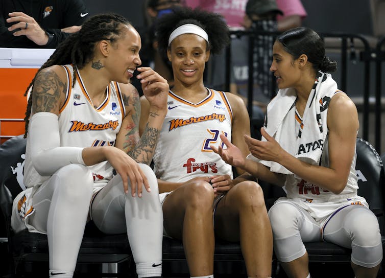 Three women basketball players are seen sitting on a bench and smiling about an amusing conversation