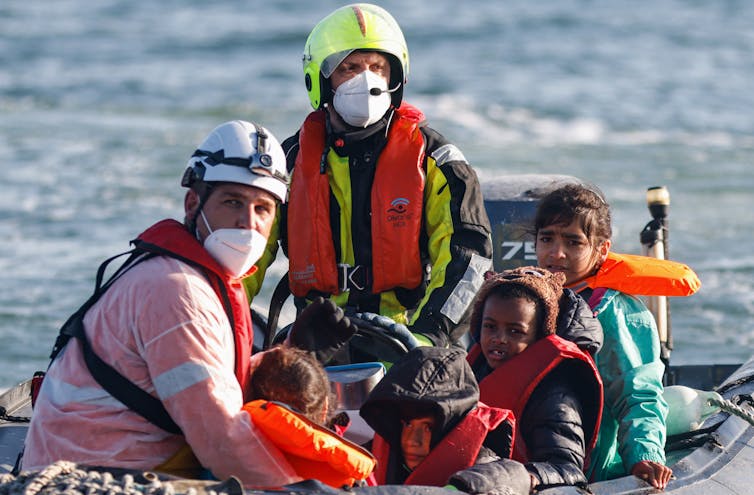 Two men wearing life jackets and masks sit with small children in a motorboat at sea.