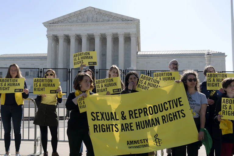 Women in front of the Supreme Court hold yellow signs that say "Sexual & reproductive rights are human rights."