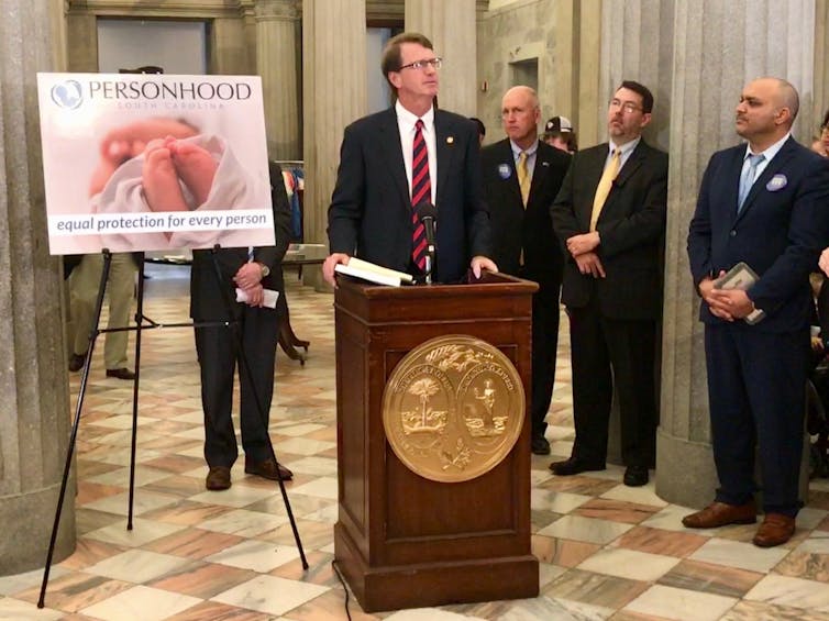 Men in suits speak in front of a poster that says