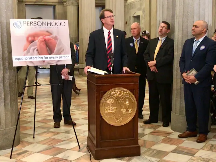 Men in suits speak in front of a poster that says