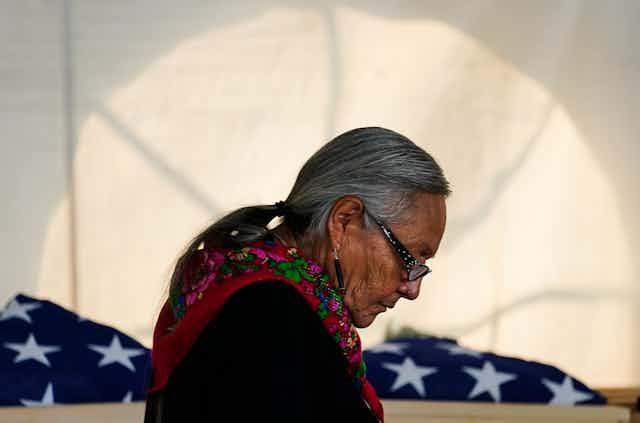 A silver-haired woman wearing a colorful scarf and glasses bows her head near folded American flags.