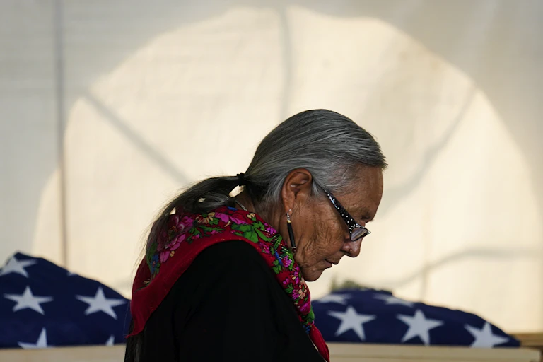 A silver-haired woman wearing a colorful scarf and glasses bows her head near folded American flags.