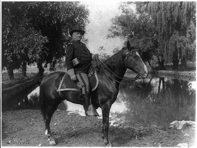 在一张明显旧照片中，一个穿着深色夹克和帽子的男人骑在一匹马上。