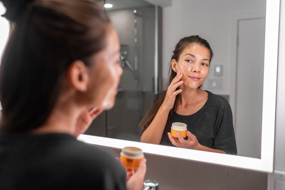 A young woman applies an anti-ageing skincare product to her face in the bathroom mirror.