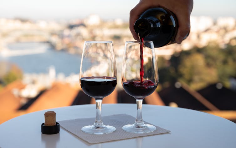Two glasses of red wine on a table with a sunny landscape in the background. Someone is pouring from the bottle into one of the glasses.
