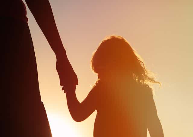 Silhouette of a young girl at sunset holding the hand of an adult, who is out of frame