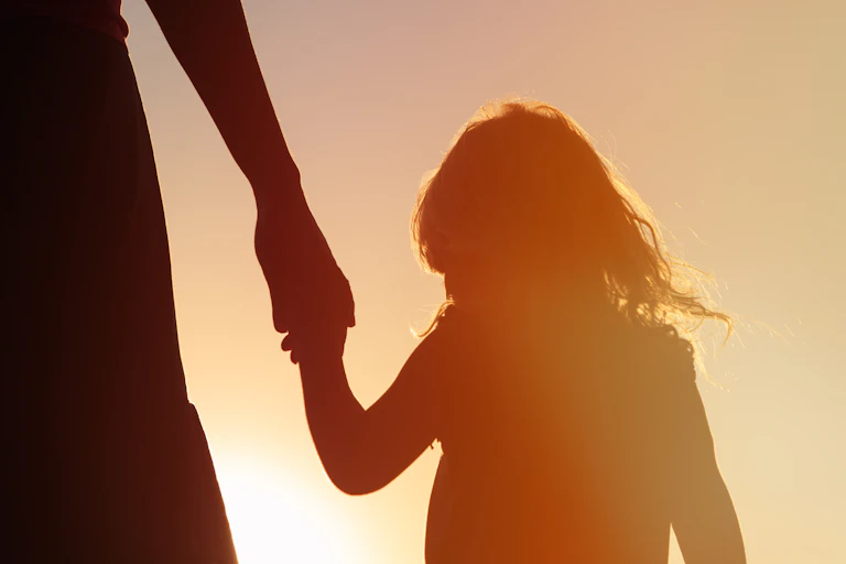 Silhouette of a young girl at sunset holding the hand of an adult, who is out of frame