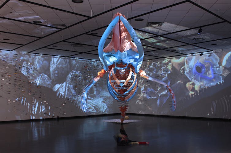 A whale skeleton made of plastic is displayed hanging in a room with images projected around it