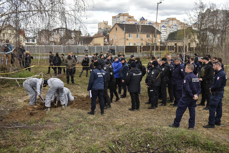Forensic investigators wearing protective clothing exhuming a mass grave in Bucha watched by officials.