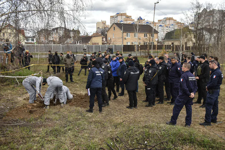 Forensic investigators wearing protective clothing exhuming a mass grave in Bucha watched by officials.