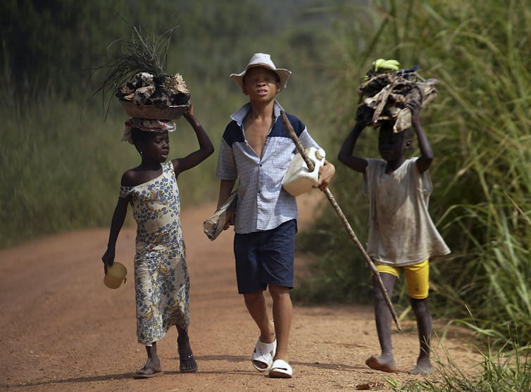 Three children walking on a dirt road carrying wood and other things on their heads.
