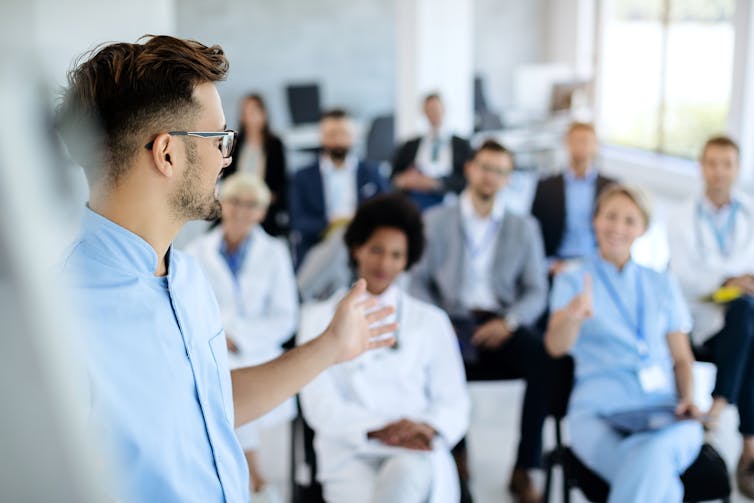 A man standing in front of a group of seated people wearing white coats and surgical scrubs