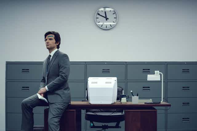 A man wearing a grey suit with a bandaid on his head sits on the edge of a desk, there is a clock in the background and the desk has an old computer on top of it.